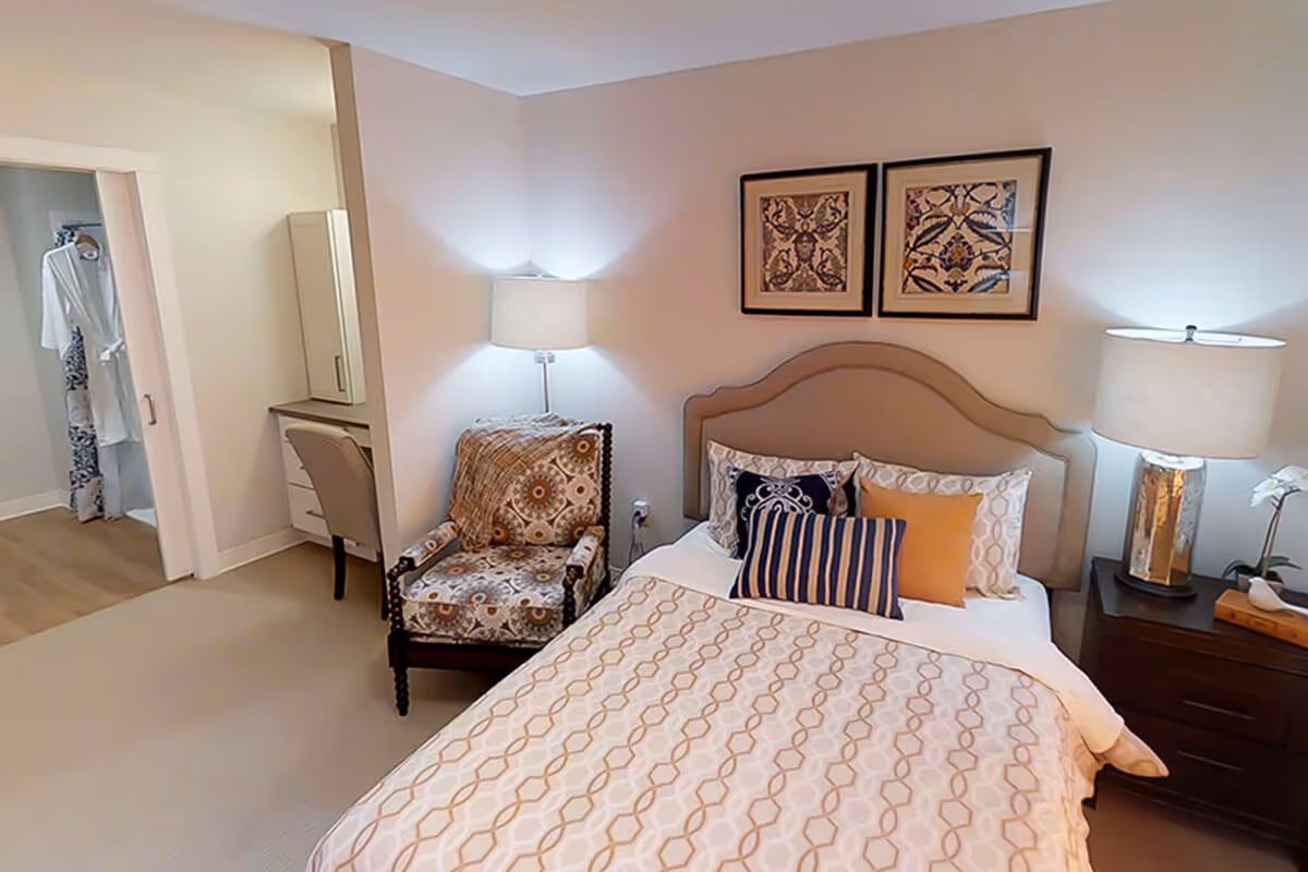 A cozy bedroom with a bed featuring patterned bedding and multiple pillows. There is a cushioned armchair with a decorative throw blanket next to a small desk and chair. Two framed artworks hang above the bed, and two lamps provide soft lighting on either side of the bed. A walk-in closet with hanging robes is visible through an open doorway.