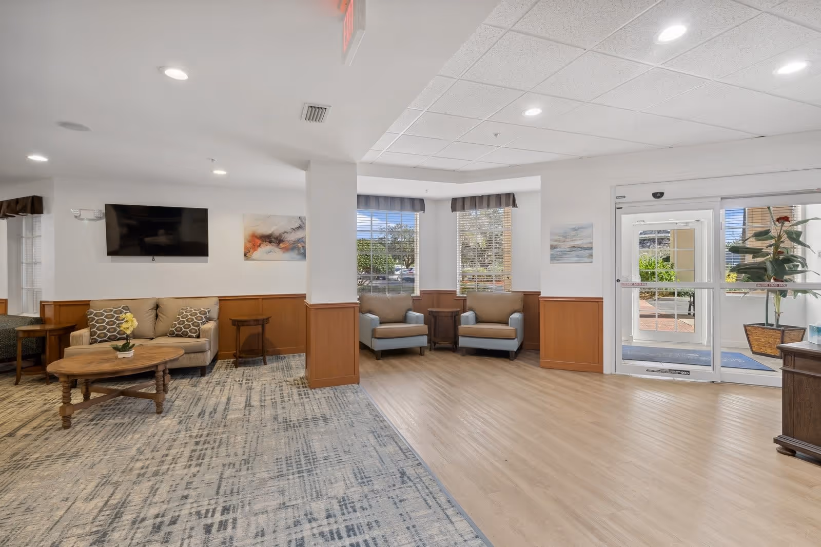 A bright and spacious senior living facility common area with a mix of carpet and wood flooring. The room features a beige loveseat with patterned pillows, two beige armchairs with a small wooden table between them, a wall-mounted flat-screen TV, and abstract wall art. Large windows with blinds allow natural light to fill the space. A glass door leads outside, and there are plants and additional furniture visible near the entrance.