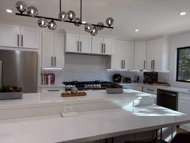 Modern kitchen with white cabinetry, a stainless steel refrigerator, stove with range hood, and a large white marble countertop island with built-in electrical outlets. A contemporary light fixture with multiple spherical bulbs hangs above the island. There are small plants and kitchen appliances on the counters, and a window on the right side lets in natural light.