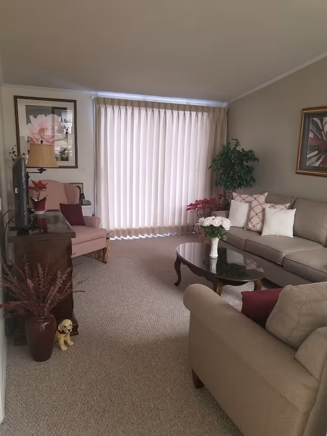 A cozy living room with beige carpet and neutral-colored walls. The room features a beige sofa with patterned and plain cushions, a glass-top wooden coffee table with a vase of white flowers, a pink armchair with a burgundy pillow, a wooden TV stand with a flat-screen TV, and various plants and framed artwork on the walls. Light beige curtains cover a large window, allowing soft natural light to enter the room.