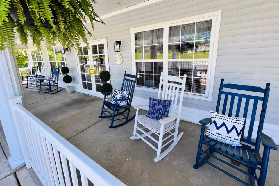 A covered porch area with several rocking chairs in blue and white, each with decorative pillows. The porch has white railings, potted topiary plants, hanging ferns, and large windows with white frames on the building's exterior wall.