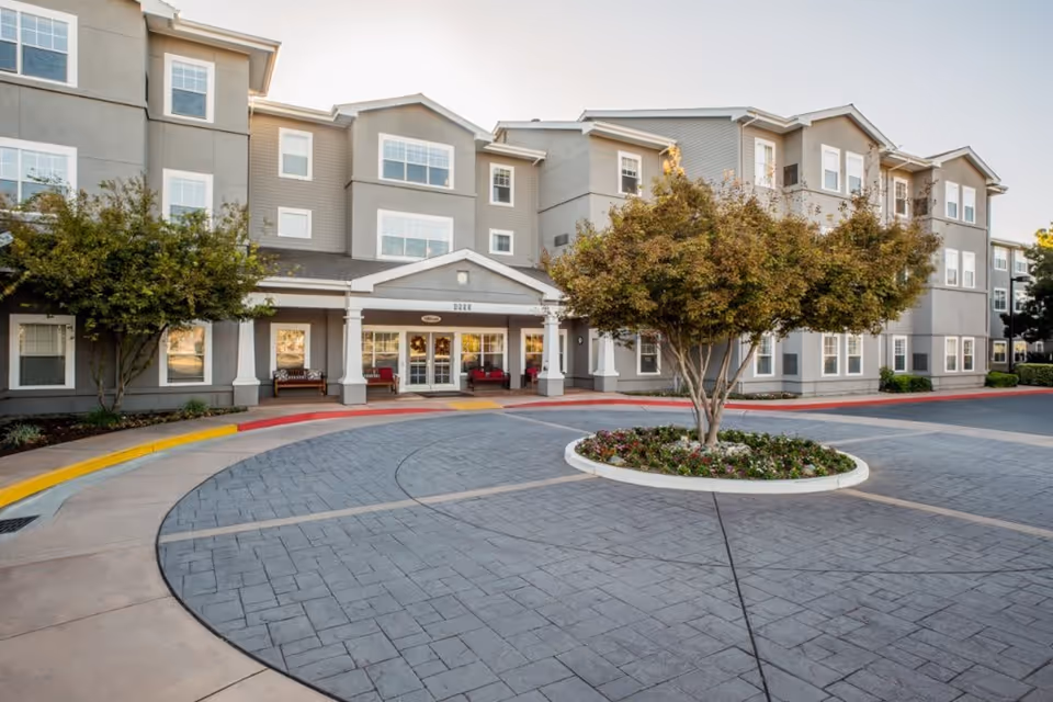 Exterior view of a multi-story senior living facility building with a circular driveway and a landscaped roundabout featuring a tree and flowers in front of the entrance.