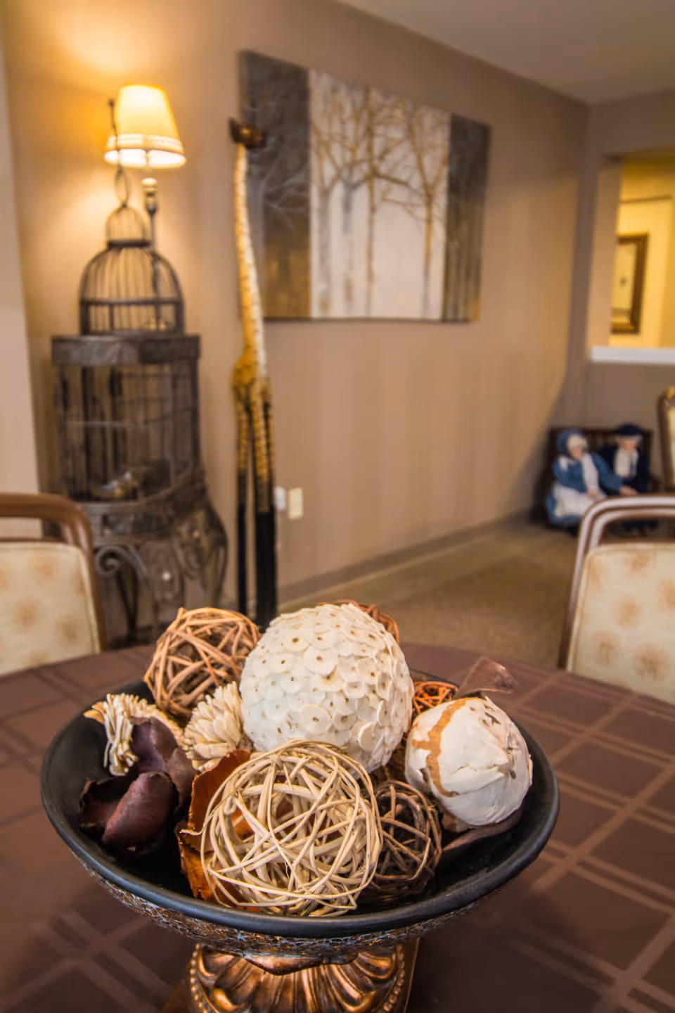 A decorative bowl of woven and dried ornamental balls sits on a table with a lamp, wall art, and chairs in a warm interior space.