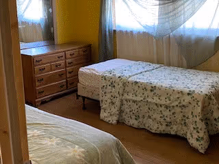 A bedroom with two single beds covered in floral bedspreads. There is a wooden dresser with multiple drawers against the wall, and two windows with sheer curtains allowing natural light into the room.