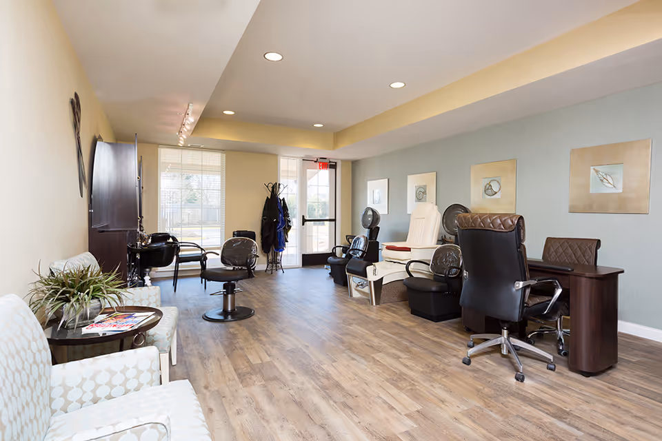 Interior view of a salon area in an assisted living facility with salon chairs, hair drying stations, a manicure table with chairs, and a seating area with two patterned armchairs and a small table with magazines. The room has wood flooring, light-colored walls, and framed artwork on the wall.