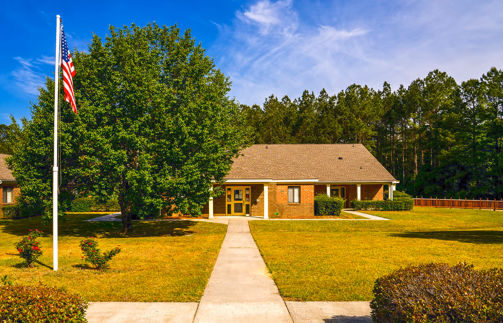 A single-story brick building with a gray shingled roof set in a grassy yard with a concrete walkway leading to the entrance. There is a large leafy tree to the left of the walkway and an American flag on a flagpole in front of the tree. The background shows a dense line of tall pine trees under a blue sky with some wispy clouds.