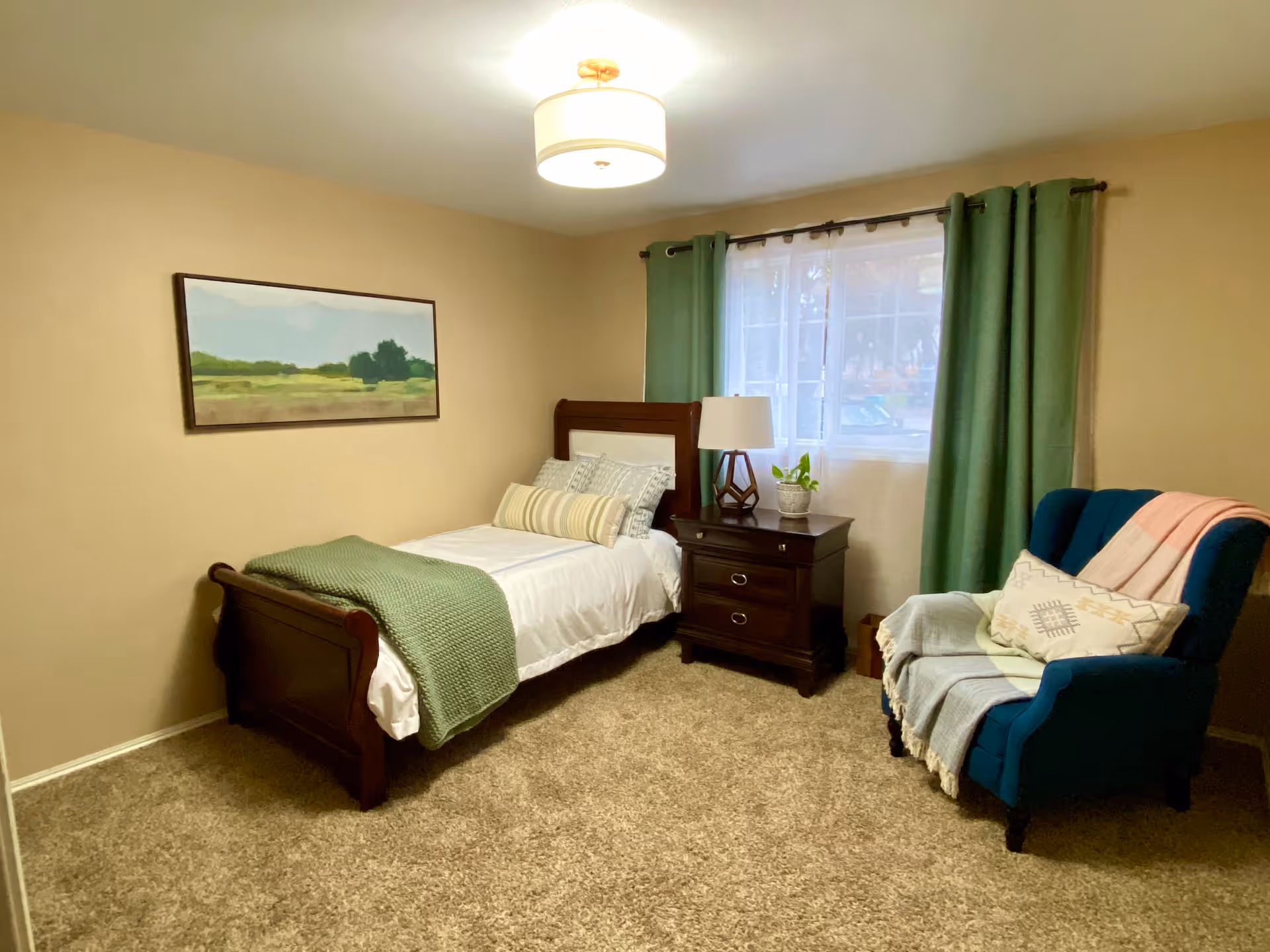 A cozy bedroom with beige walls and carpeted floor. The room features a single wooden bed with white bedding, a green throw blanket, and decorative pillows. Next to the bed is a dark wooden nightstand with a lamp and a small potted plant. A large window with green curtains and sheer white drapes lets in natural light. A blue upholstered armchair with a pink and a light blue throw blanket and a decorative pillow is positioned near the window. A landscape painting hangs on the wall above the bed.