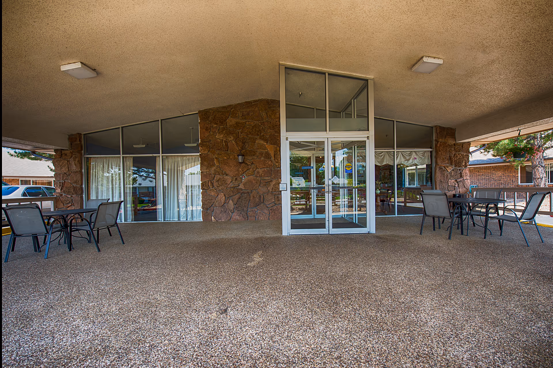 Covered entrance area of a building with glass double doors in the center, stone walls on either side, and large windows with curtains. There are two outdoor seating areas with tables and chairs on both sides of the entrance. The ground is covered with a textured surface.