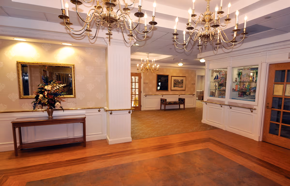 An elegant interior hallway of Oak Knoll Healthcare Center featuring polished wooden floors, beige patterned wallpaper, white wainscoting, and brass handrails. The space is decorated with chandeliers hanging from the ceiling, a wooden table with a floral arrangement, framed artwork on the walls, and a glass display case with decorative items. A wooden door with glass panels is visible on the right side.