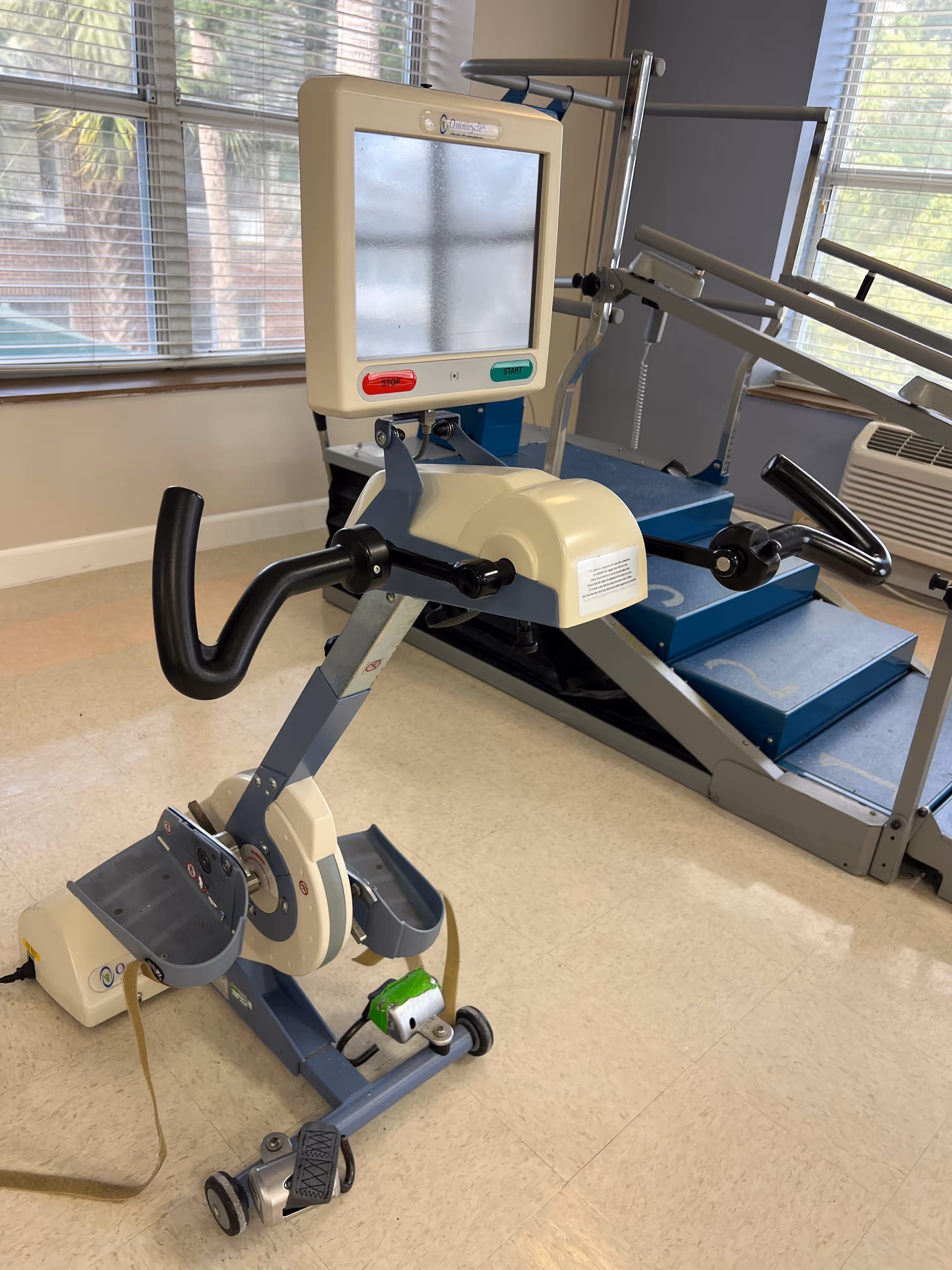 A physical therapy exercise machine with pedals and handlebars, positioned in a room with large windows and a blue rehabilitation stair stepper in the background.