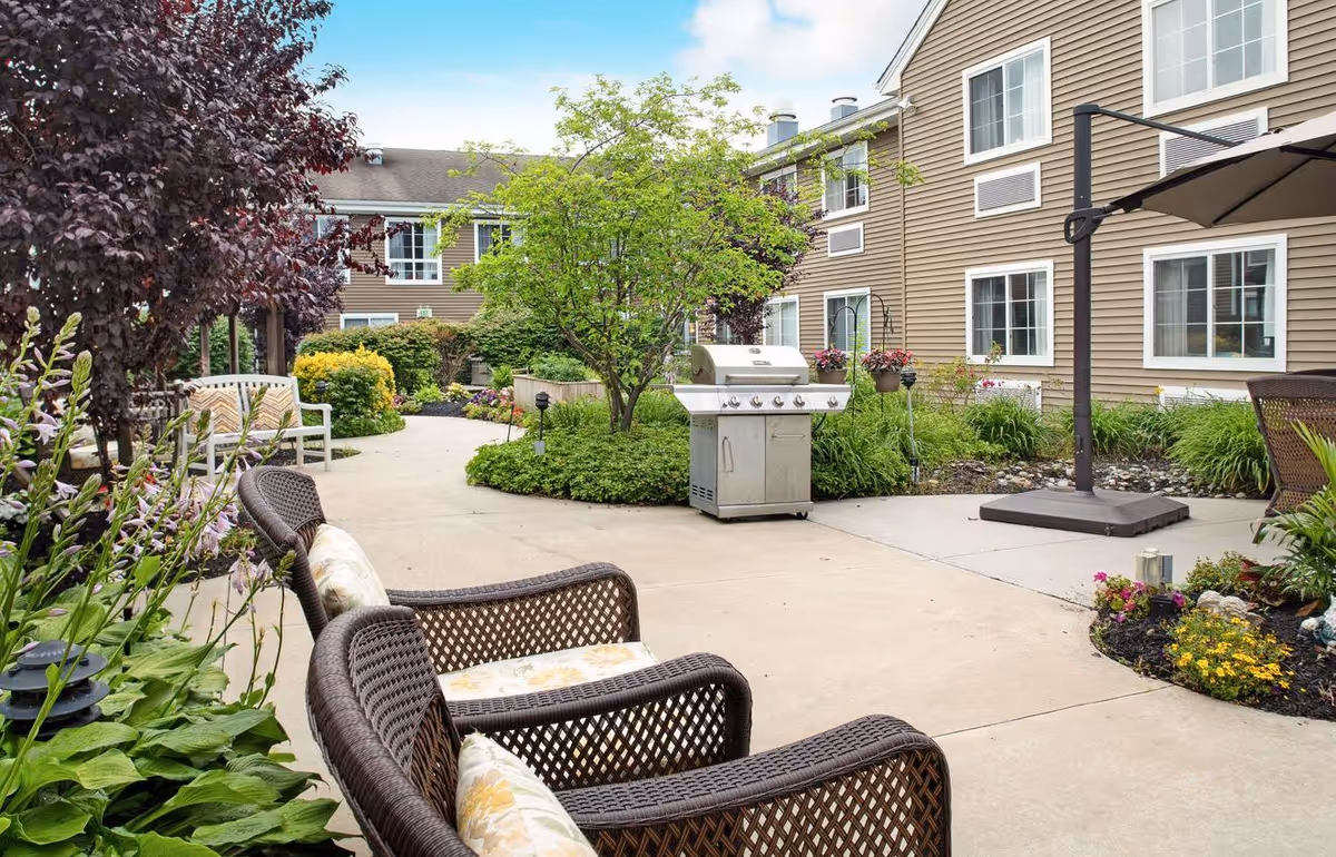 Outdoor patio area at Brighton Gardens of Edison featuring wicker chairs with cushions, a stainless steel grill, a large umbrella, and well-maintained garden beds with various plants and flowers surrounding a paved walkway. The building exterior with multiple windows is visible in the background.