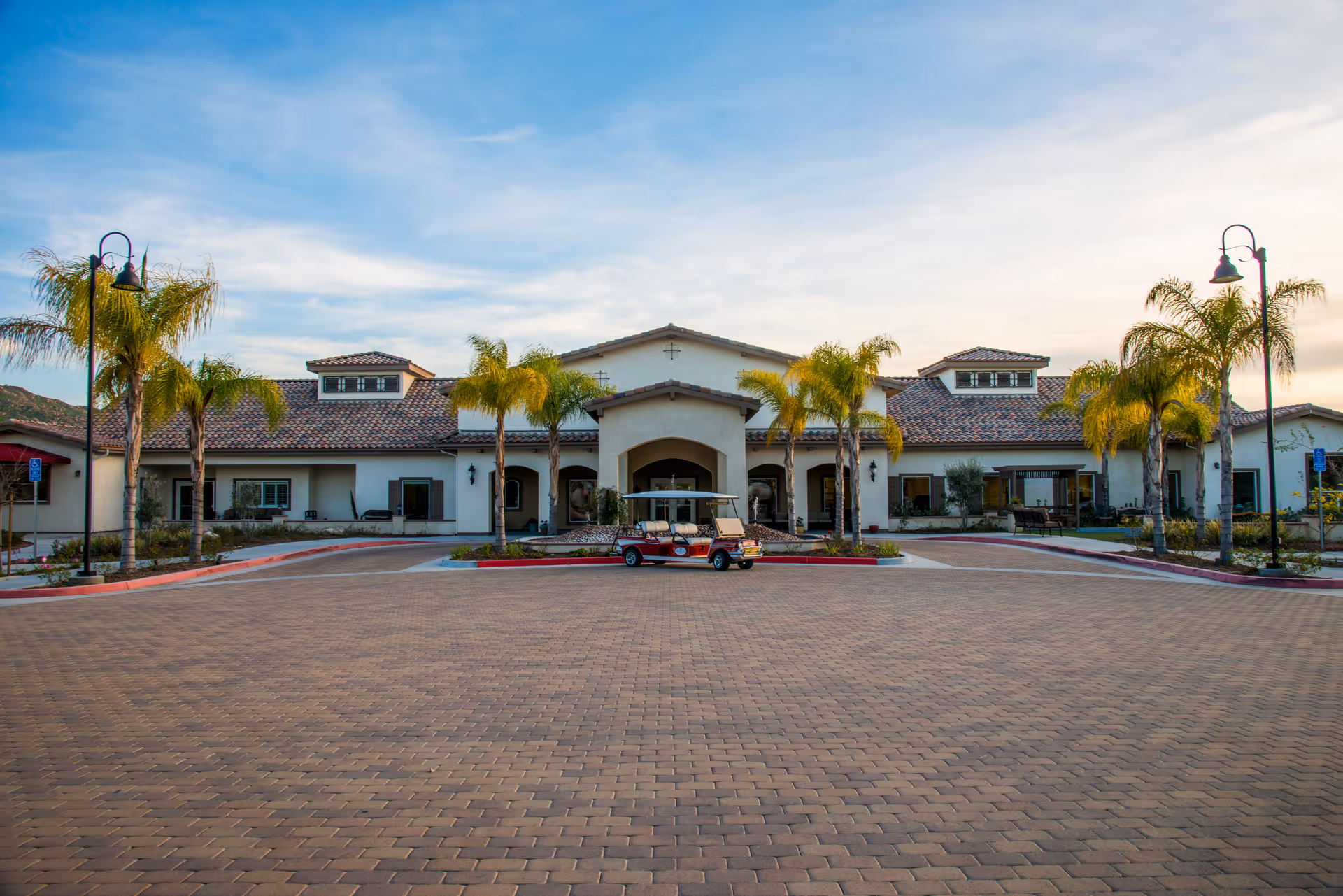 Front entrance of a single-story Mediterranean-style senior living building with palm trees and a small golf cart parked in the circular driveway.