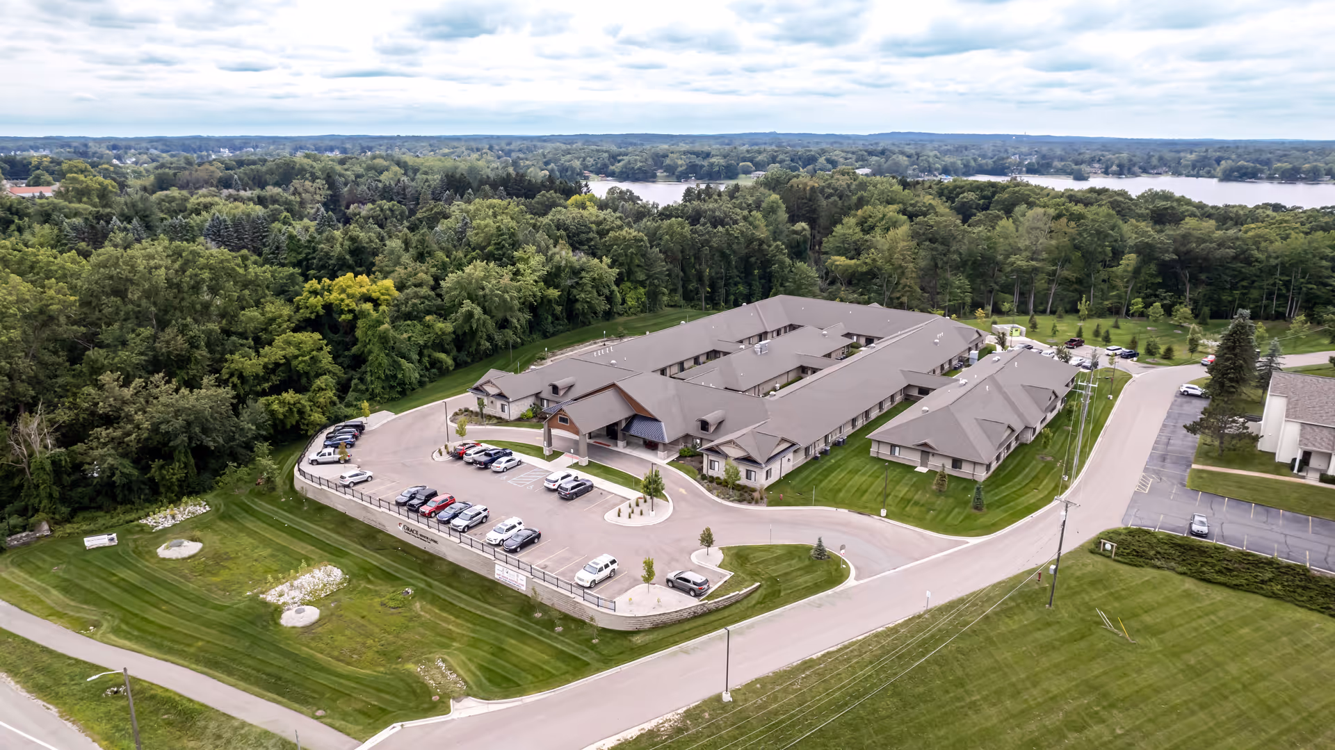 Aerial view of a single-story senior living campus with connected buildings, a parking lot, landscaped lawns, surrounding trees, and a lake in the distance.
