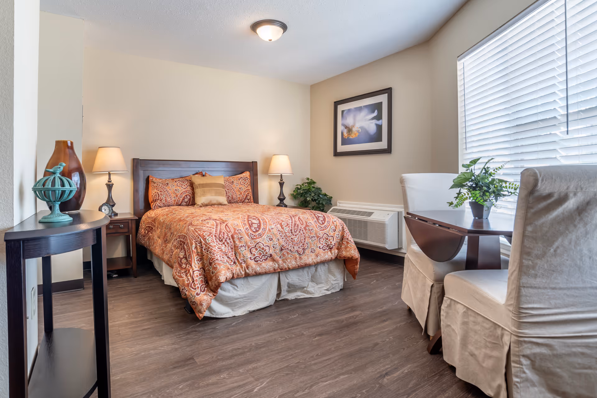 A cozy bedroom with a double bed covered in an orange patterned comforter, two bedside tables with lamps, a framed flower picture on the wall, a small table with two chairs near a window with blinds, and decorative plants.