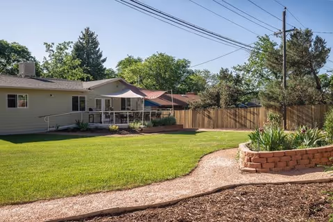 A spacious backyard area of a single-story building with a well-maintained green lawn, a gravel pathway, a raised flower bed with plants, and a wooden fence. There are trees and power lines visible in the background, and a covered patio attached to the building.