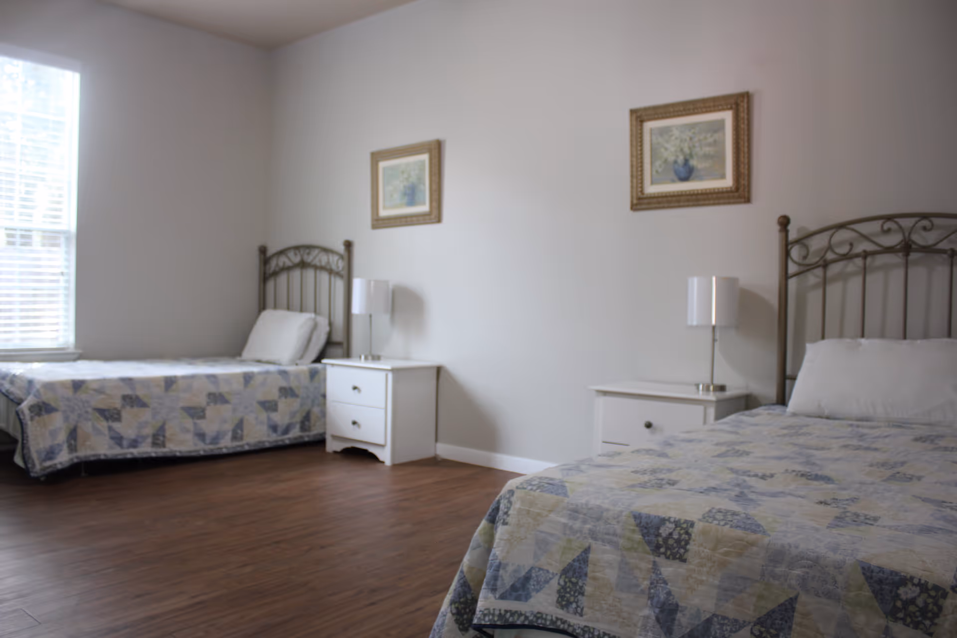 A simple, clean bedroom with two twin beds, each with a metal headboard and a patterned quilt. Between the beds are two white nightstands, each with a white lamp. Two framed floral paintings hang on the light gray wall above the nightstands. A window with blinds is on the left side, allowing natural light to enter the room. The floor is wooden.