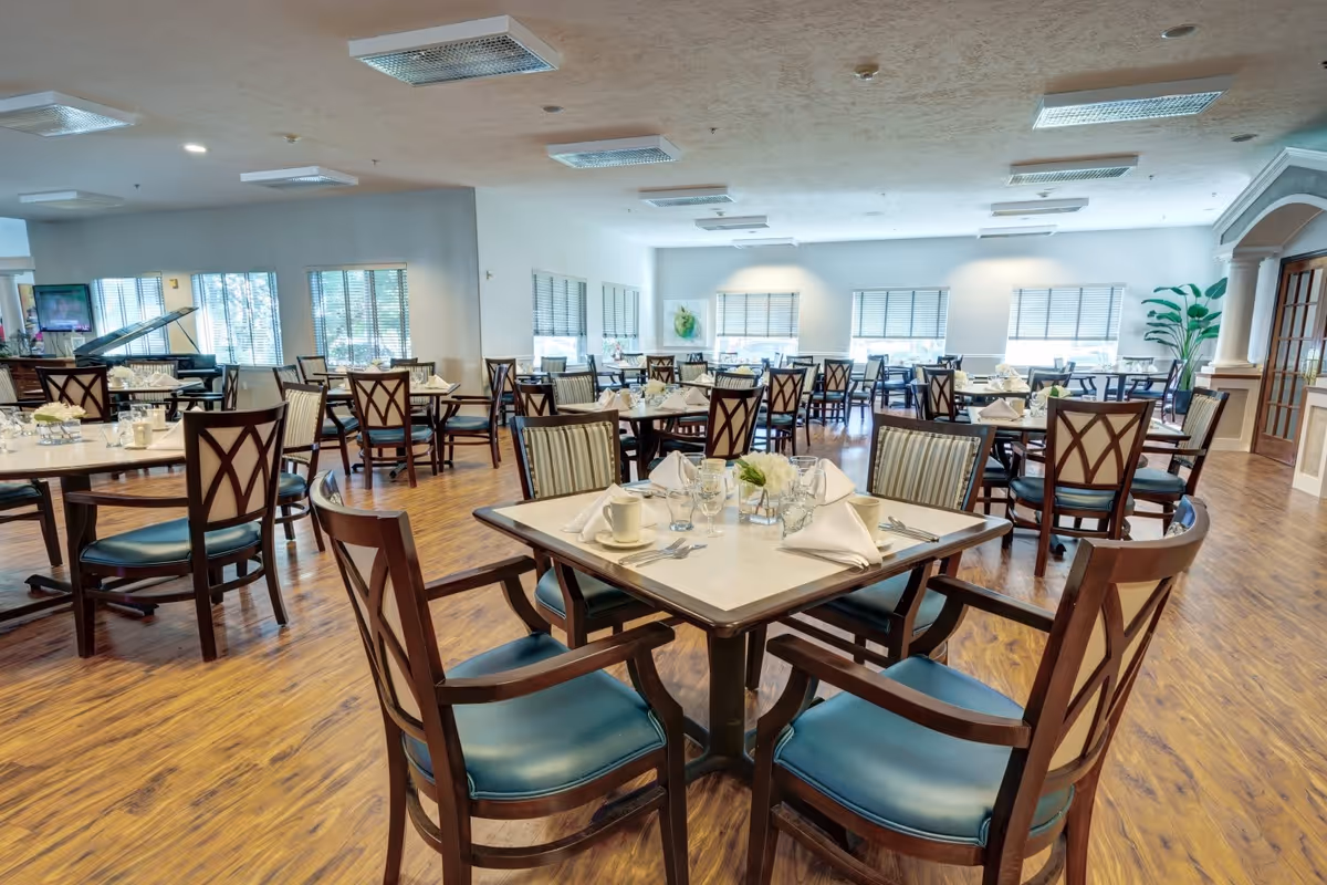 A spacious dining room with multiple square tables set with white napkins, glassware, and small floral centerpieces. The room features wooden floors, large windows with blinds, and wooden chairs with blue cushions. A grand piano is visible in the background near the windows.