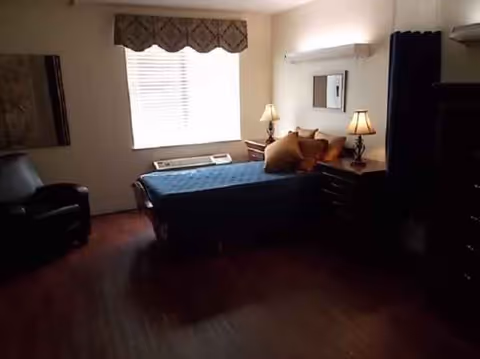 Dimly lit bedroom in a senior living facility with a bed, lamps, window, and dresser.