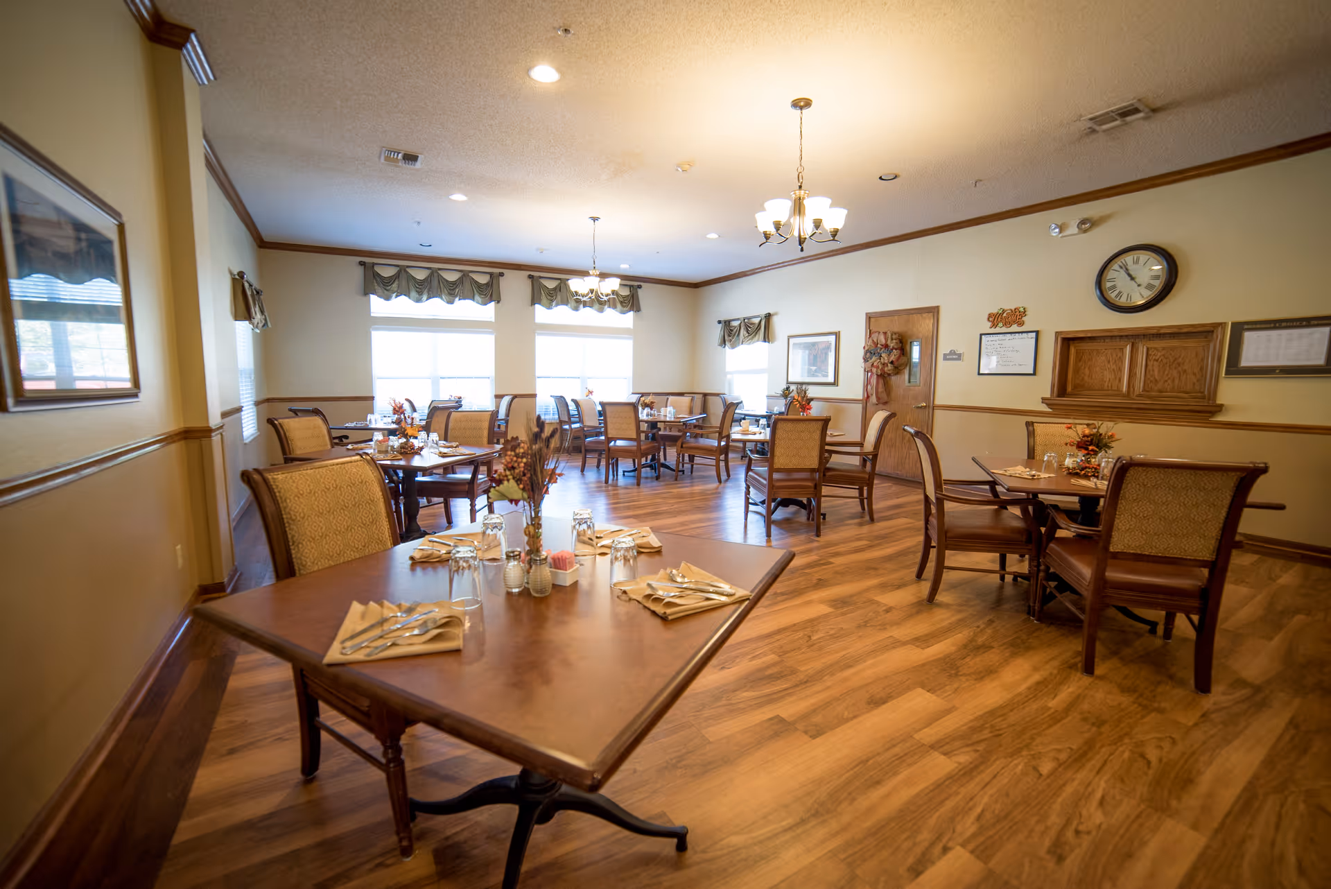 A bright, neatly arranged dining room with wooden tables and chairs set for meals and large windows.