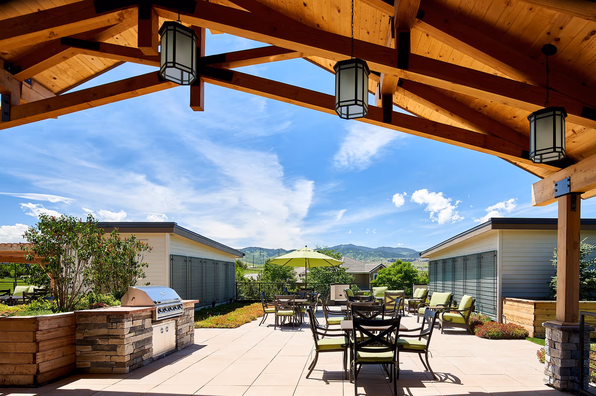 Outdoor patio area at MorningStar Senior Living of Arvada with a wooden pergola overhead, hanging lantern-style lights, several tables and chairs with green cushions, a green umbrella, a built-in stone grill, and a scenic view of mountains and blue sky in the background.