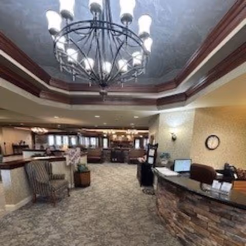 Interior view of a senior living facility lobby or common area with a large chandelier hanging from a tray ceiling, a stone-faced reception desk with a computer and office supplies, a striped armchair, and a spacious carpeted floor leading to a dining or lounge area in the background.