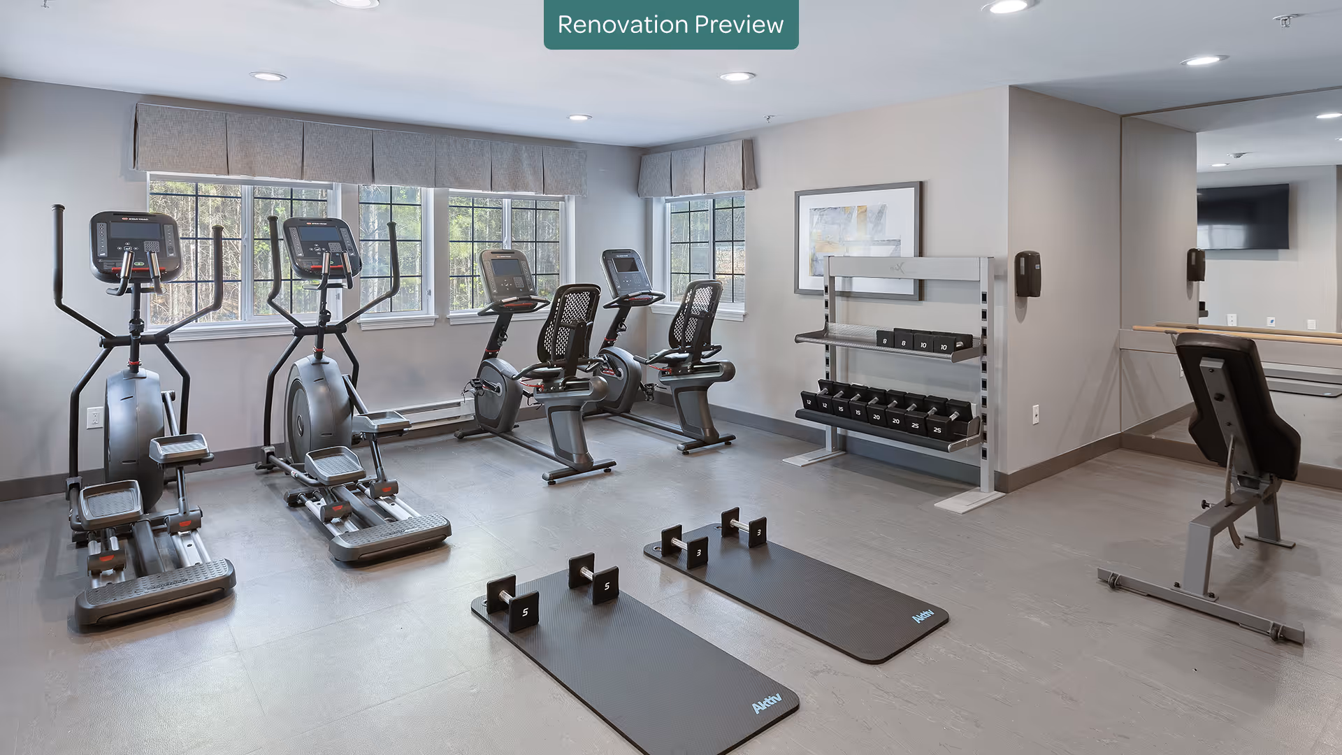 A modern fitness room with two elliptical machines, two recumbent exercise bikes, two exercise mats with dumbbells, a rack of various dumbbells, a weight bench, large windows with gray valances, and a wall mirror. The room has light gray walls and flooring, and recessed ceiling lights. A sign at the top center reads 'Renovation Preview'.