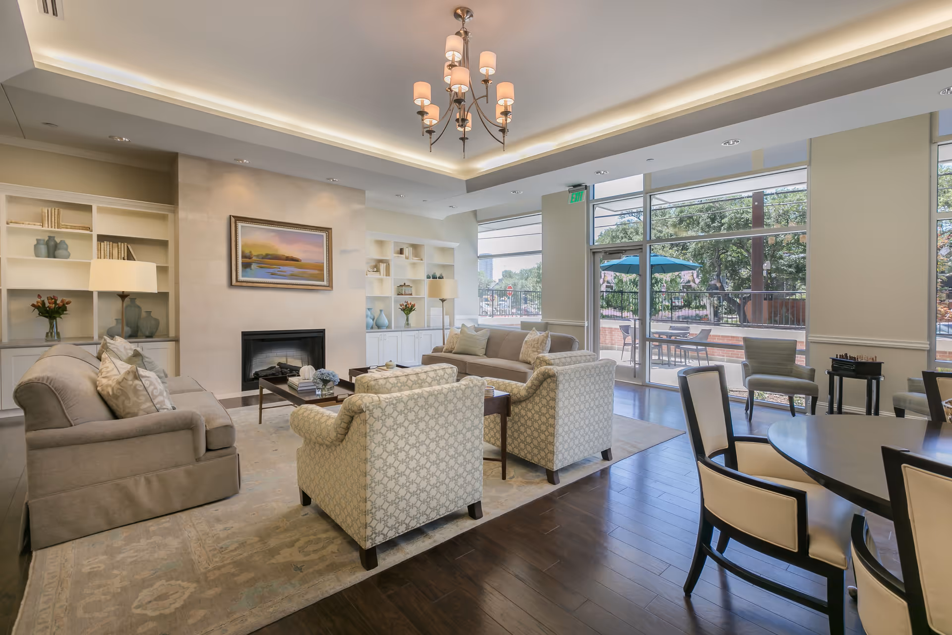 A bright and spacious living room area with comfortable sofas and armchairs arranged around a coffee table in front of a fireplace. Built-in shelves with decorative items and books flank the fireplace. Large windows and glass doors let in natural light and offer a view of an outdoor patio with tables and umbrellas. A chandelier hangs from the ceiling, and there is a round dining table with chairs in the foreground.