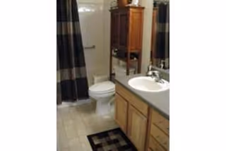 Bathroom with a toilet, a sink with a countertop and wooden cabinets, a shower with a curtain, and a small rug on the tiled floor.