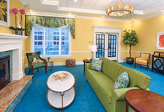 A bright and inviting living room with a green sofa adorned with patterned pillows, a round white coffee table, two armchairs, a fireplace with a marble hearth, a large window with floral curtains, and a chandelier hanging from a coffered ceiling. The walls are painted yellow and the carpet is blue.