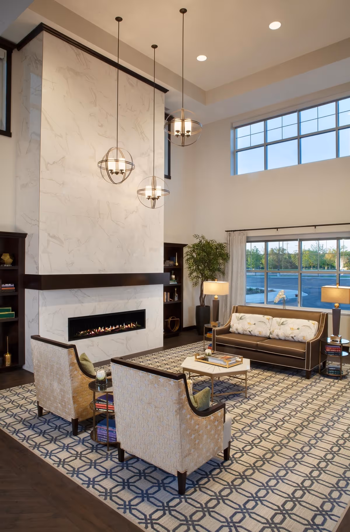 A modern living room with a tall white marble fireplace, three spherical pendant lights hanging from the ceiling, two beige armchairs with green cushions, a brown leather sofa with floral pillows, a patterned area rug, side tables with lamps, books, and a large window showing an outdoor view.