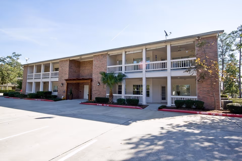 Exterior view of a two-story senior living facility building with brick and white siding, featuring balconies on the upper floor and covered walkways on the ground floor. There are small bushes and a palm tree in front, with a clear blue sky above and a parking area in front of the building.
