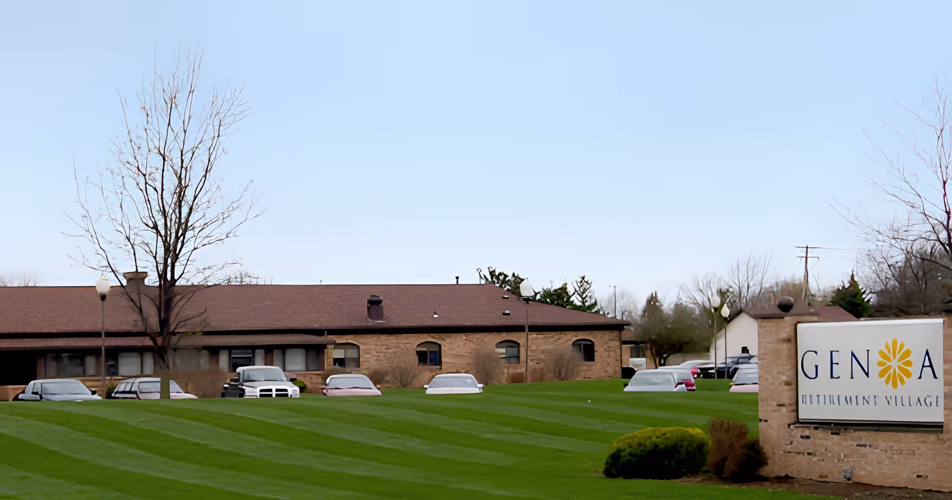 Brick senior living building and parking lot behind a striped green lawn with a Genoa Retirement Village sign on the right.