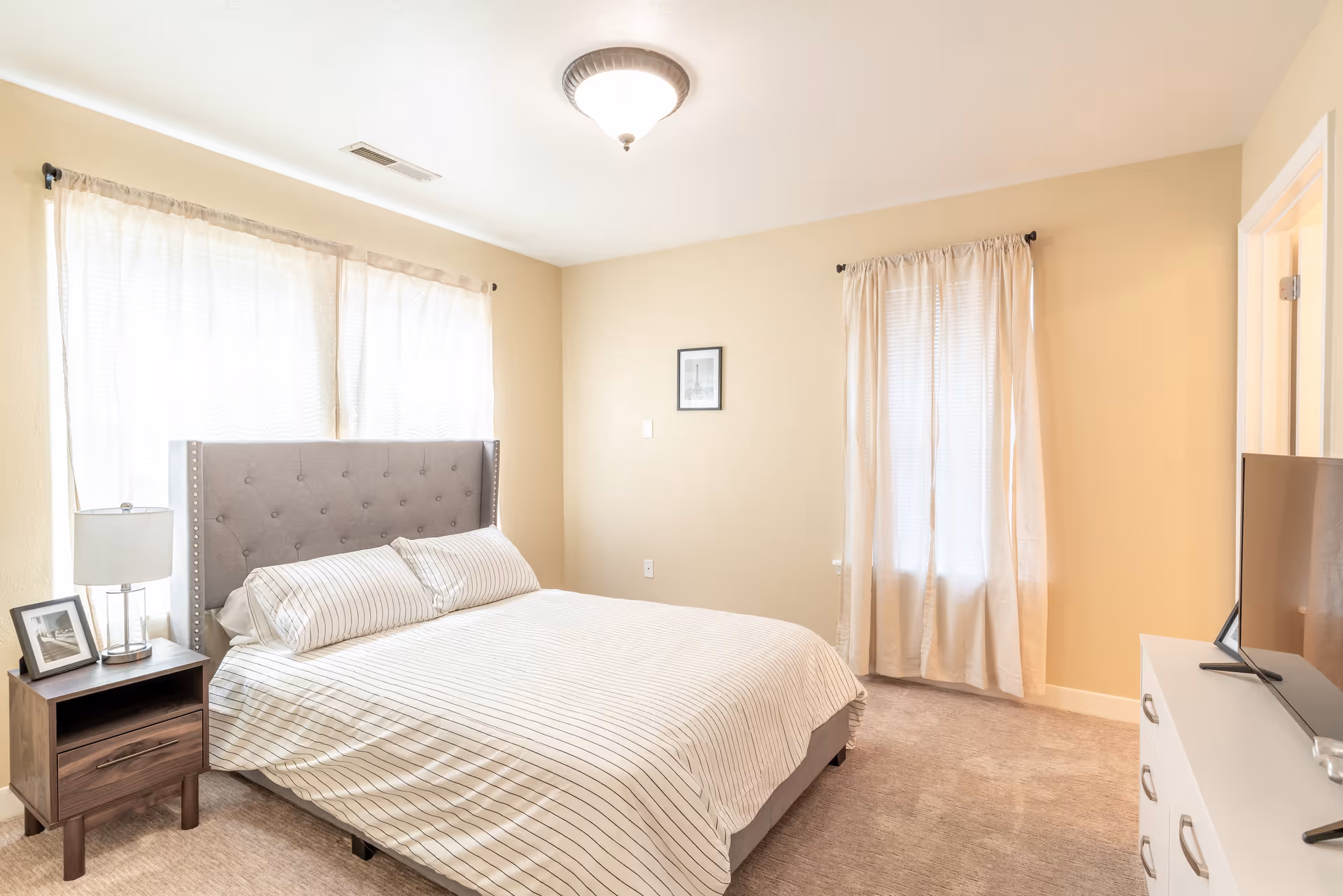 A bright bedroom with a large bed featuring a tufted gray headboard and striped bedding. There is a wooden nightstand with a lamp and framed photo on the left side of the bed. Two windows with light curtains allow natural light into the room. A white dresser with a flat-screen TV on top is positioned on the right side of the room.