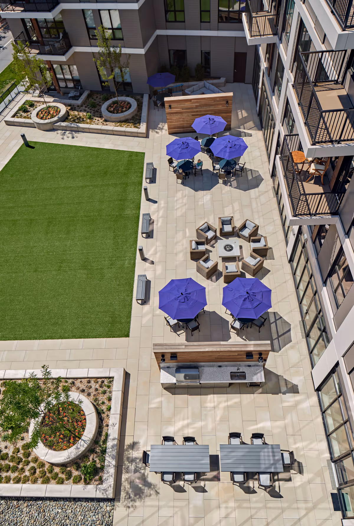Aerial view of an outdoor courtyard area at a senior living facility featuring a large green lawn, several seating areas with purple umbrellas, a circular fire pit surrounded by chairs, picnic tables with chairs, and a built-in outdoor kitchen with a grill. The courtyard is bordered by a multi-story building with balconies overlooking the space and landscaped garden beds.