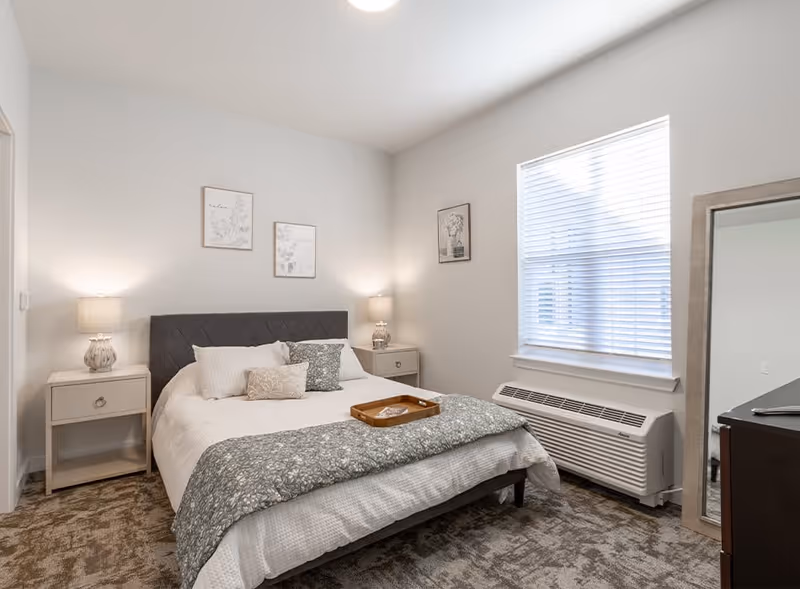 A neatly arranged bedroom with a queen-sized bed featuring a dark upholstered headboard, white bedding, and decorative pillows. Two bedside tables with matching lamps flank the bed. There are three framed artworks on the walls. A large window with blinds allows natural light into the room. A full-length mirror leans against the wall near a dark dresser. The floor is carpeted with a patterned design.