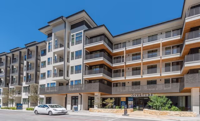 Exterior view of a modern multi-story senior living facility named Overture Domain with balconies, large windows, and a clear blue sky. A white car is parked in front of the building.