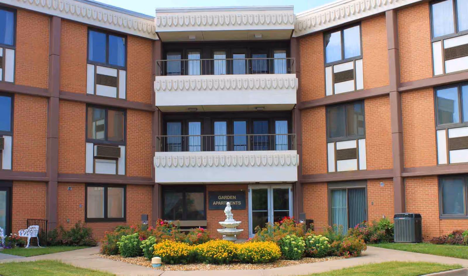 Exterior view of a three-story brick apartment building with balconies and large windows. In front of the building is a garden area with colorful flowers and a white fountain. There is a sign that reads 'Garden Apartments' near the entrance.