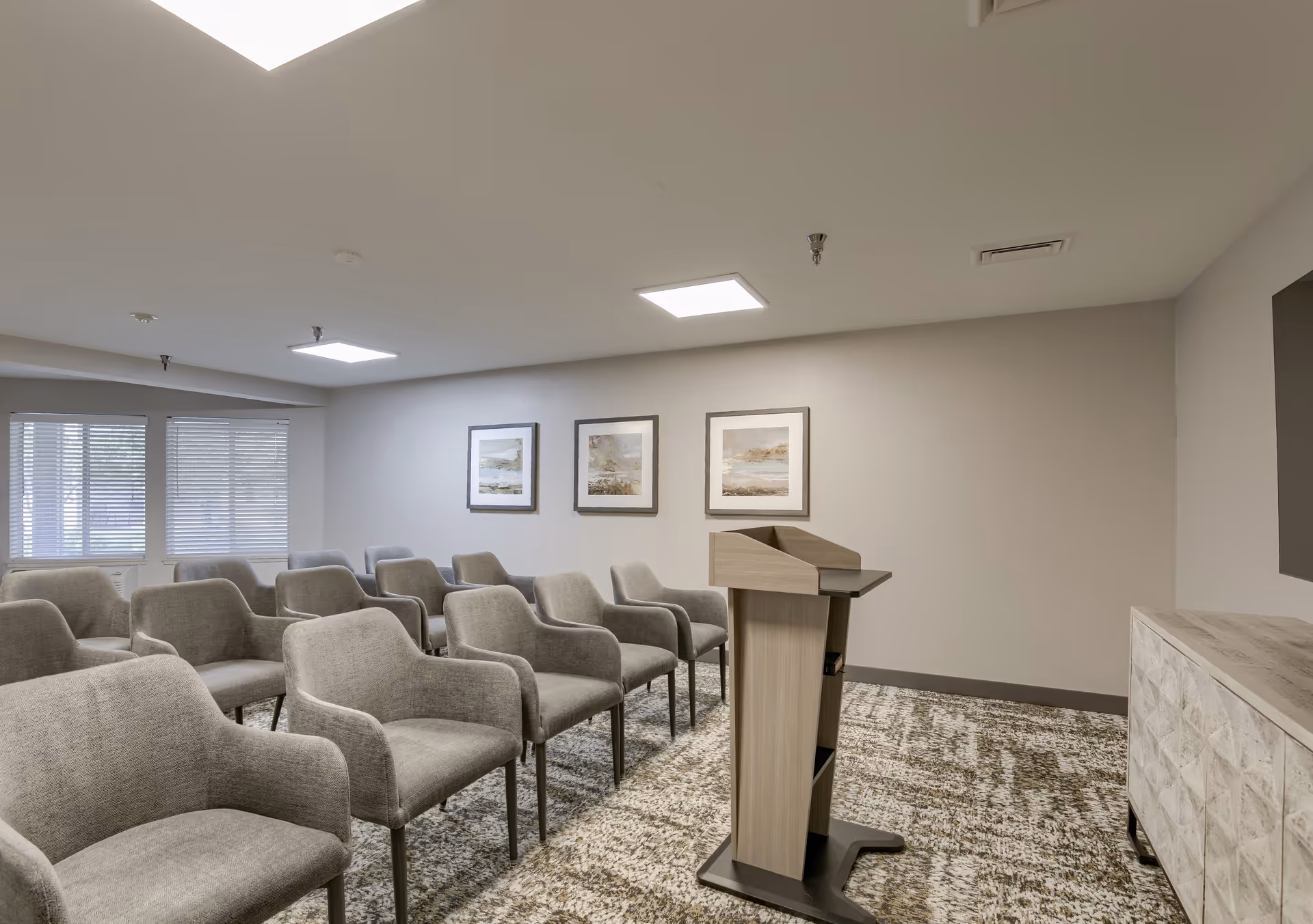 A small conference or meeting room with rows of gray upholstered chairs facing a wooden podium. The room has a patterned carpet, three framed landscape paintings on the wall, and windows with blinds letting in natural light.