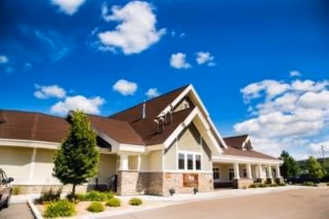 Front exterior of a single-story memory care building with peaked roofs, stone accents, landscaping, and a driveway under a blue sky.