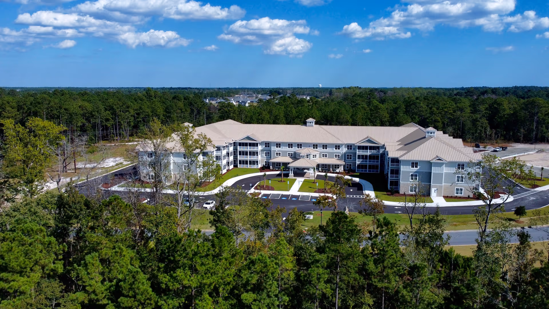 Aerial view of a large senior living facility building surrounded by trees and greenery under a blue sky with scattered clouds. The building has a beige roof and light-colored exterior walls, with a parking area and pathways in front.