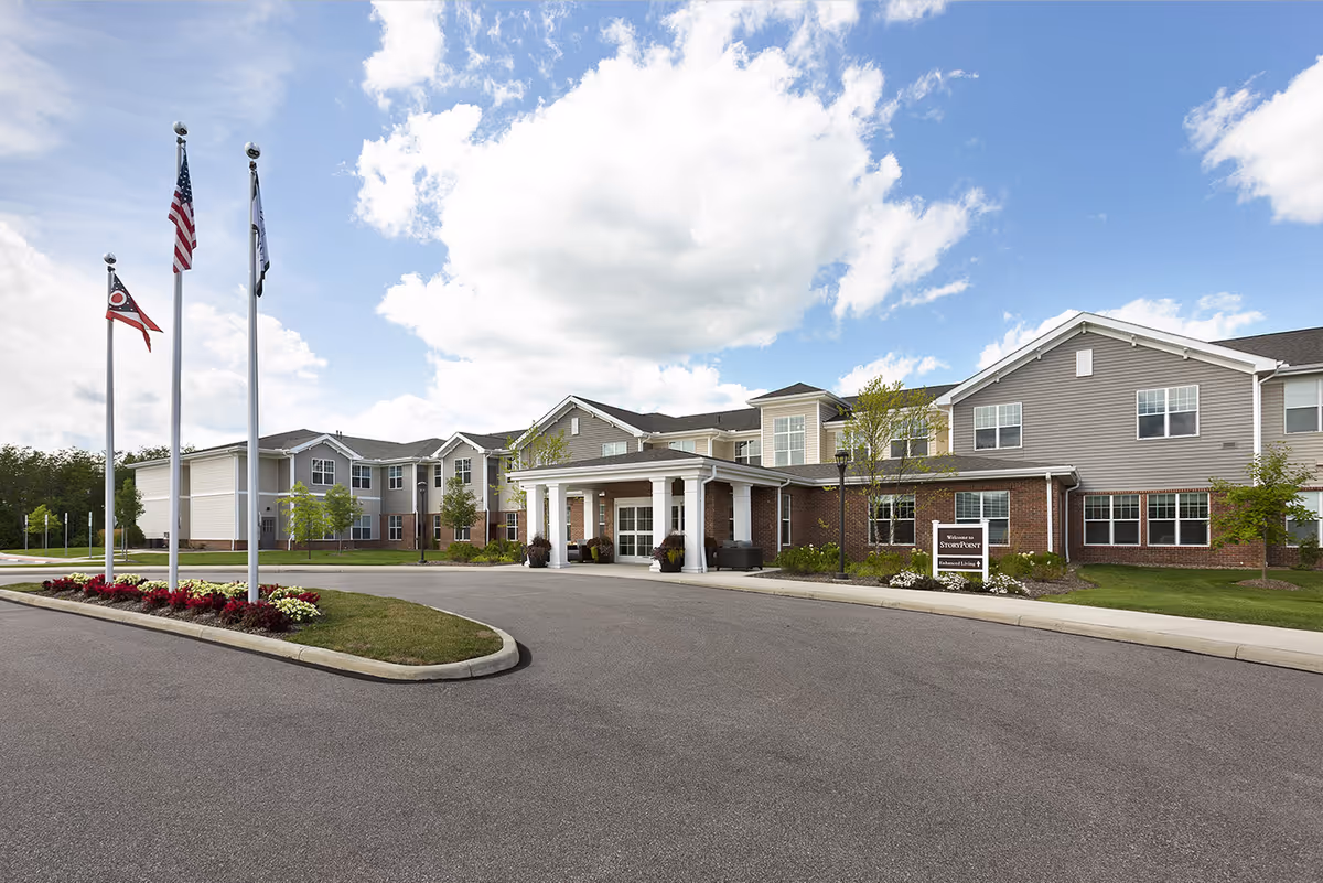 Front exterior of the StoryPoint Medina senior living building with a covered entrance, flagpoles, and landscaped drive under a partly cloudy sky.