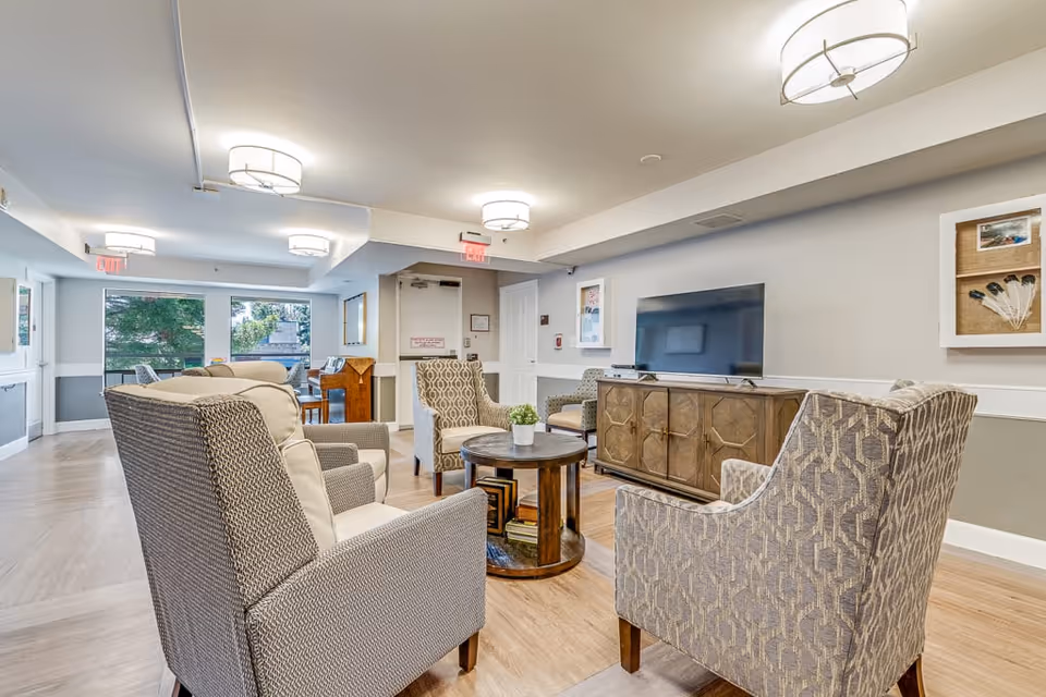 A bright and spacious senior living common area with several upholstered armchairs arranged around a round wooden coffee table. A flat-screen TV is mounted on a wooden cabinet against the wall. Large windows let in natural light, and there is a piano in the background near the windows. The room has light-colored walls and wood flooring, with modern ceiling lights.