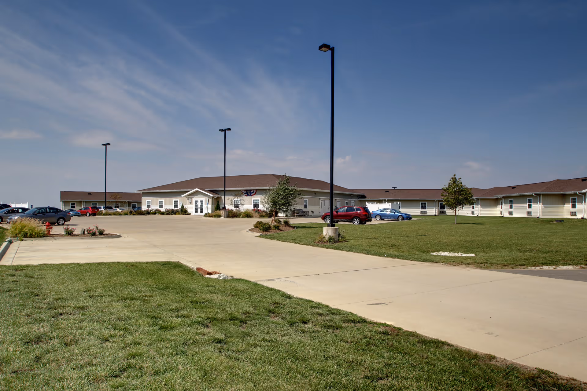 Single-story senior care facility building with a parking lot, lawn, and lamp posts under a blue sky.