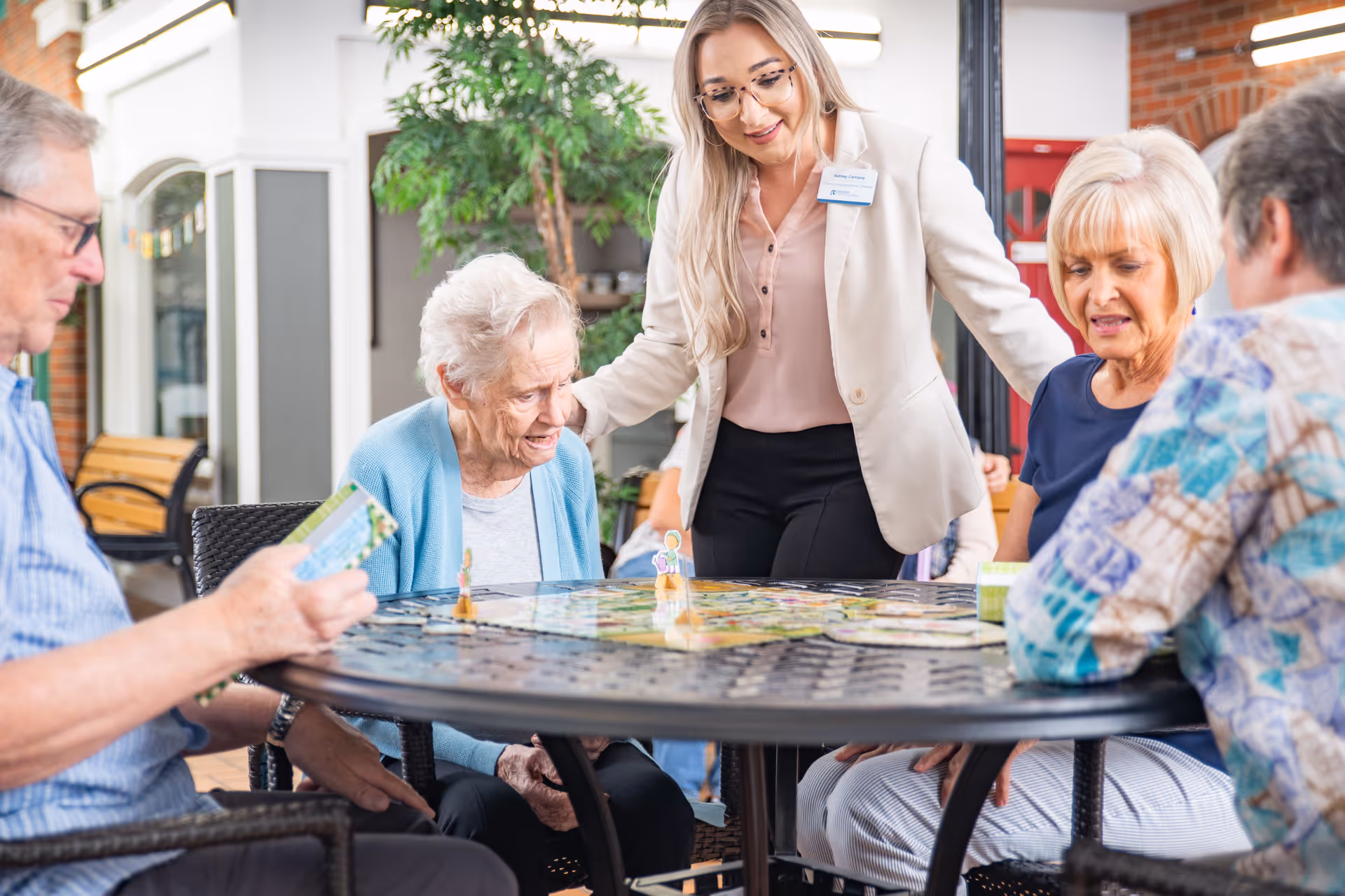 A group of elderly people sitting around a round table playing a board game, accompanied by a smiling caregiver standing and engaging with them in a bright indoor common area with plants and brick walls.