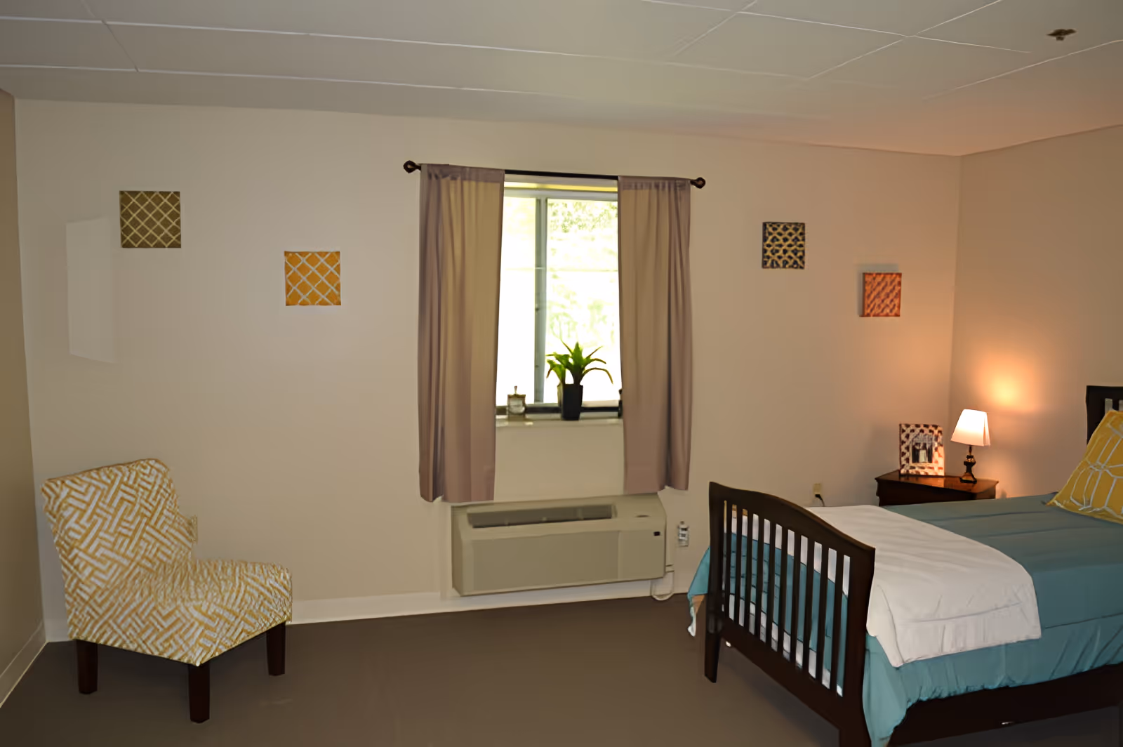 A cozy bedroom with a single bed featuring teal bedding, a white blanket, and a yellow pillow. Next to the bed is a wooden nightstand with a small lamp and a framed photo. A window with beige curtains and a potted plant on the windowsill is centered on the wall. On the left side of the room, there is a patterned yellow and white armchair. The walls are decorated with four small square art pieces in various colors.