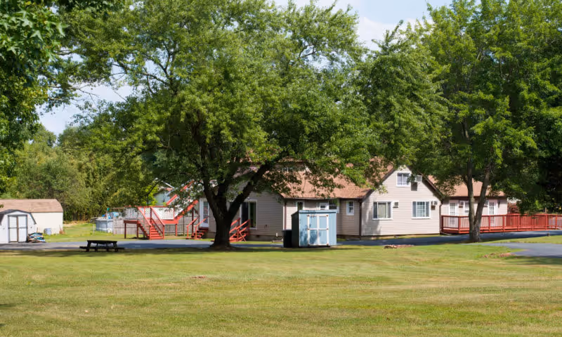 Grassy lawn and large trees in front of a single-story beige building with red decks and small storage sheds.