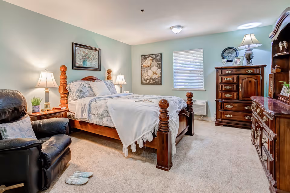 A cozy bedroom with a wooden bed frame featuring turned posts, a floral bedspread, and a white throw blanket. There are two bedside tables with lamps, a black leather recliner chair with a pillow, and a pair of slippers on the carpeted floor. The room also has a wooden dresser and chest of drawers, a window with blinds, and framed artwork on the walls.