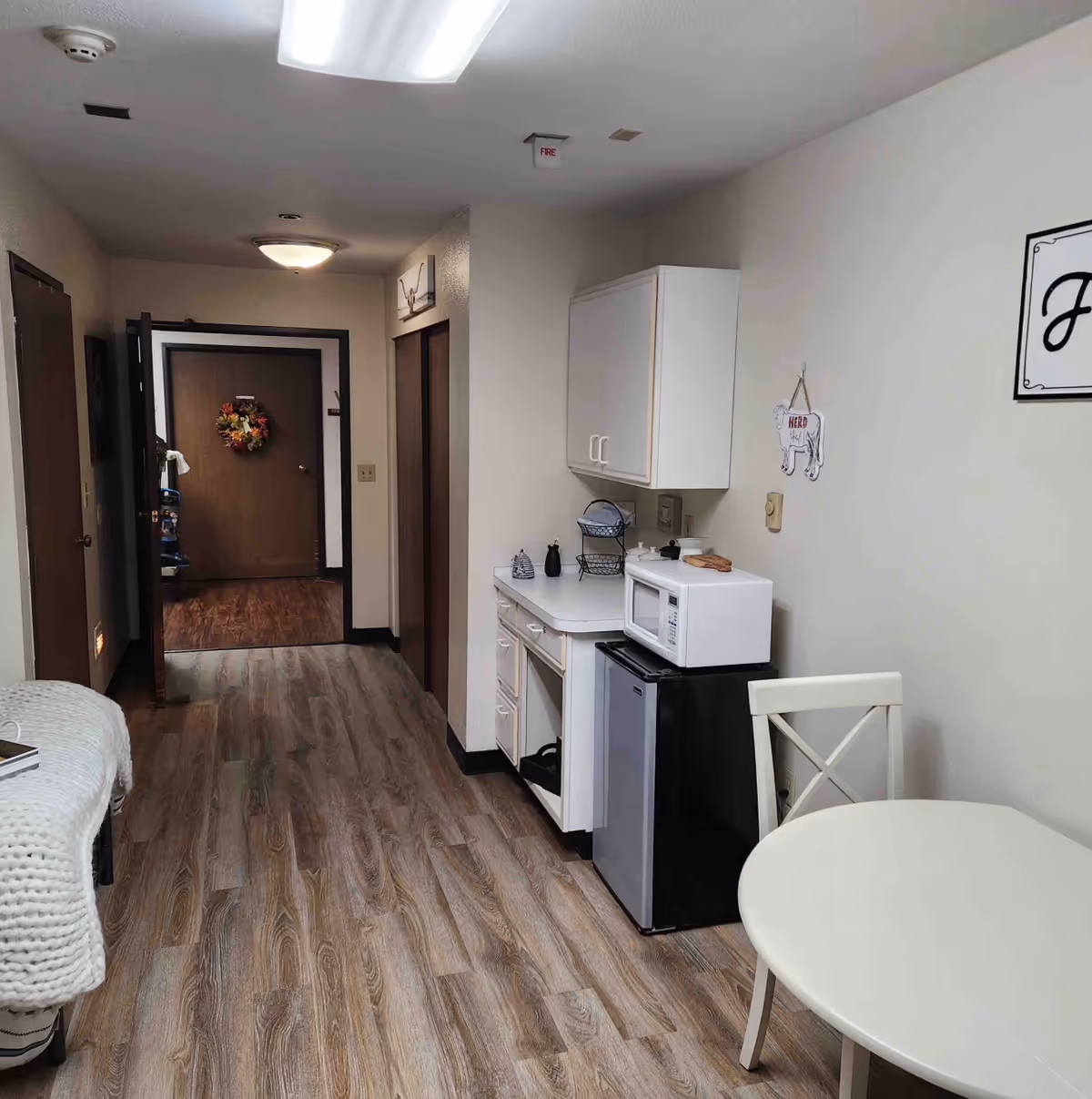 Interior view of a small kitchenette area in a senior living facility with a white round table and chair, a mini refrigerator, microwave, white cabinets, and a hallway leading to a door decorated with a wreath.