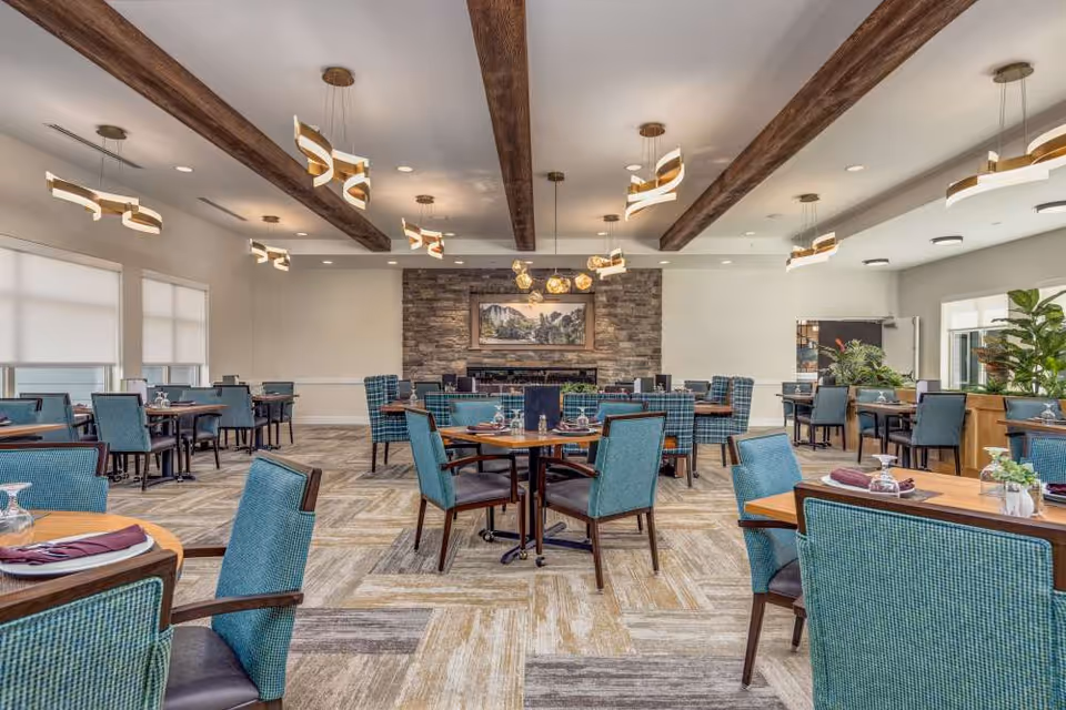 A spacious dining room with multiple wooden tables and teal upholstered chairs arranged neatly. The room features a stone accent wall with a framed landscape painting above a fireplace. Modern pendant lights hang from the ceiling with exposed wooden beams. Large windows with white blinds allow natural light to fill the room, and there are plants placed near the windows.