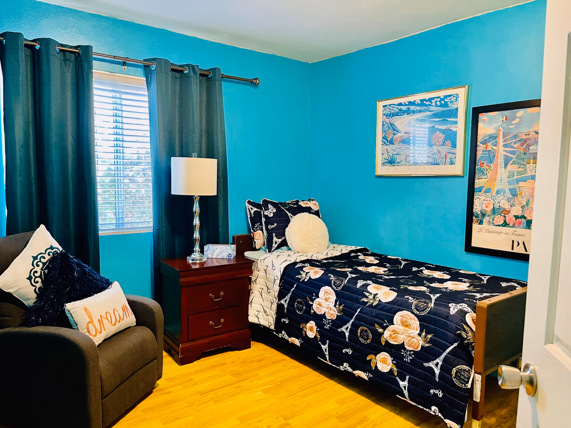 Small bedroom with a single floral bed against turquoise walls, a nightstand and lamp, an armchair with pillows, and framed artwork.